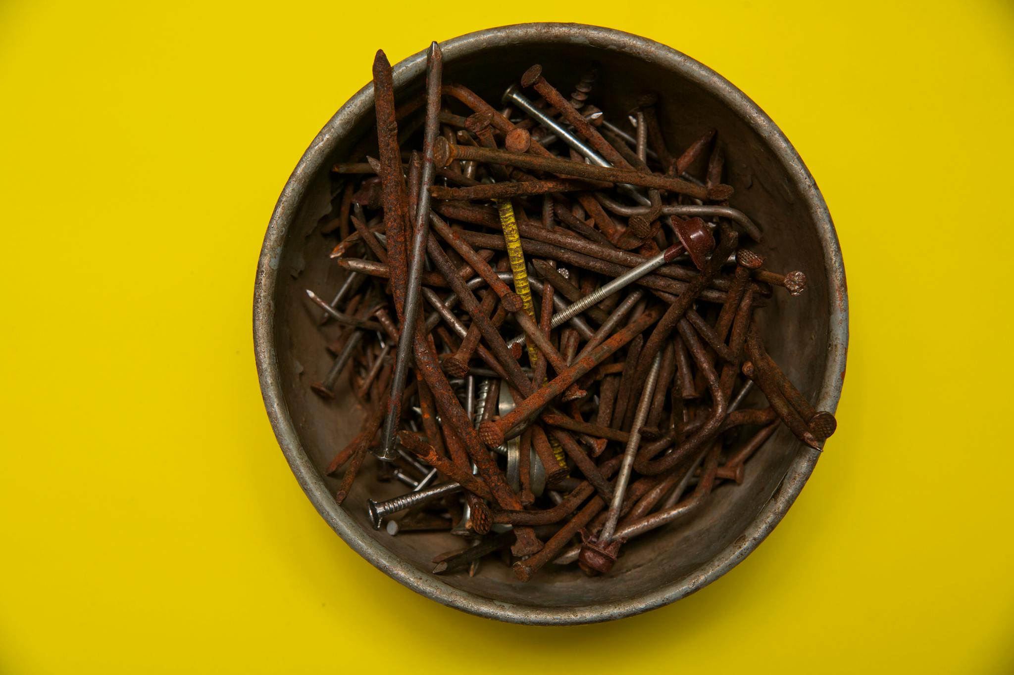 Overhead view of a metal bowl filled with rusty nails against a vibrant yellow backdrop.